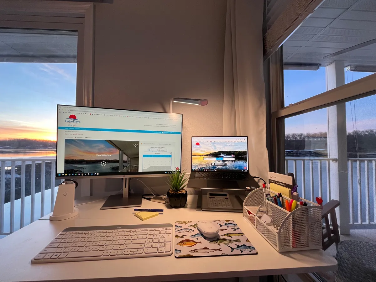 A dual-monitor desk faces a window with a colorful sunset, complemented by neat stationery and a small plant. The organized setup suggests a space designed for focused work, planning, or creative thinking, while the view adds a calm, inspiring touch.