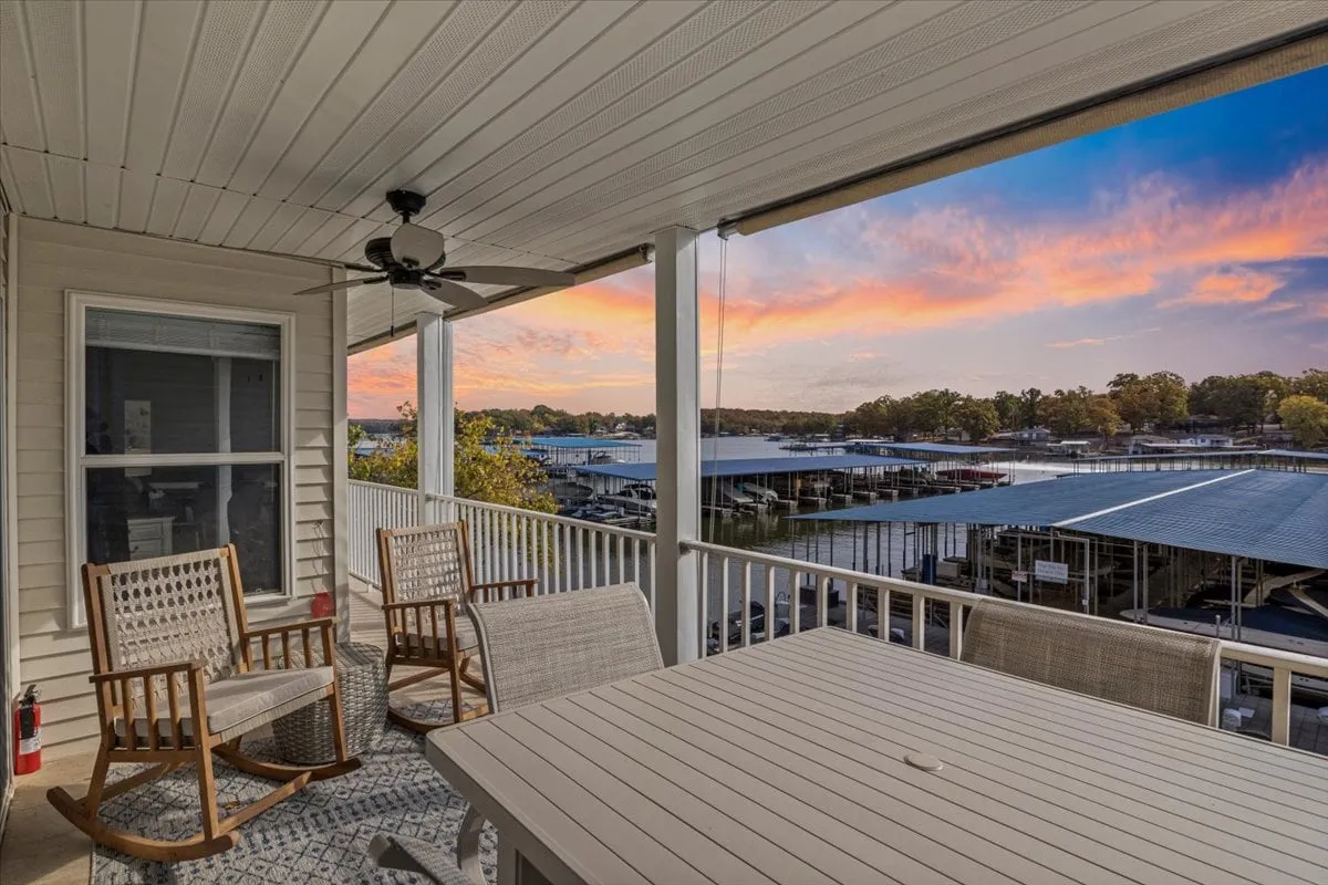 This image captures a lakeside deck furnished with rocking chairs and an outdoor table. The calm water, marina, and glowing sunset create a peaceful, inviting atmosphere that is perfect for alfresco dining and evening relaxation. The view includes boats drifting by and a sense of proximity to the water's edge.