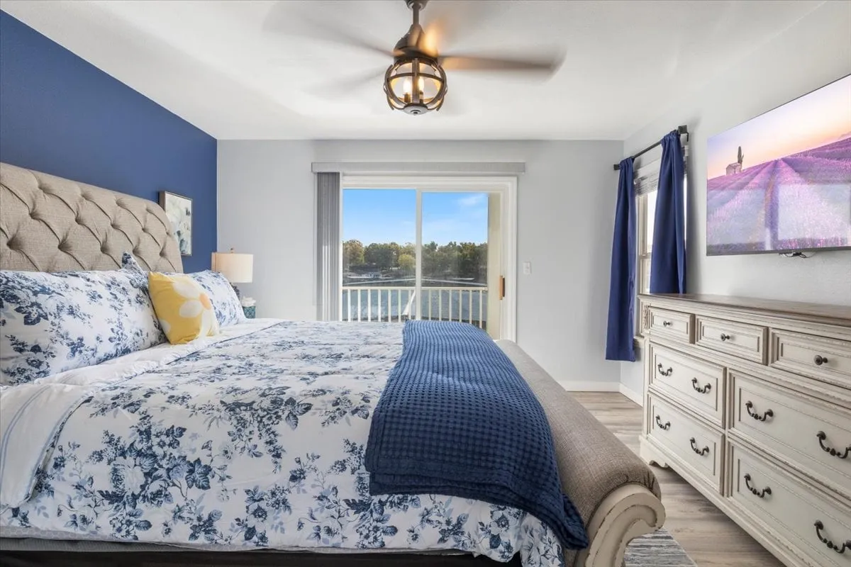 A spacious bedroom with floral bedding, a blue accent wall, a large window with a view, a dresser with decorative items, and a flat-screen TV. The room feels both homey and luxurious, perfect for couples or families.