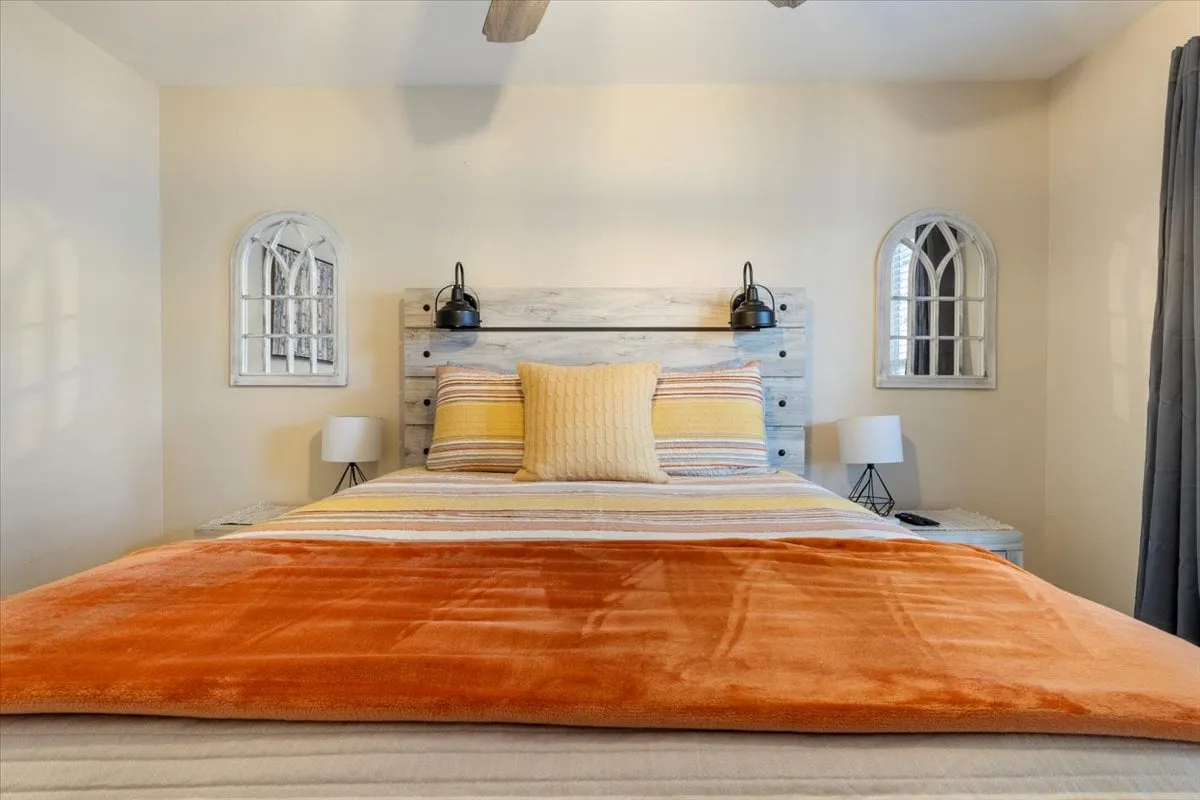 A cozy bedroom featuring a bed with colorful bedding, a wooden headboard, decorative lamps on nightstands, and arched window accents that allow natural light to filter in. The space feels warm, inviting, and tailored for rest and relaxation.