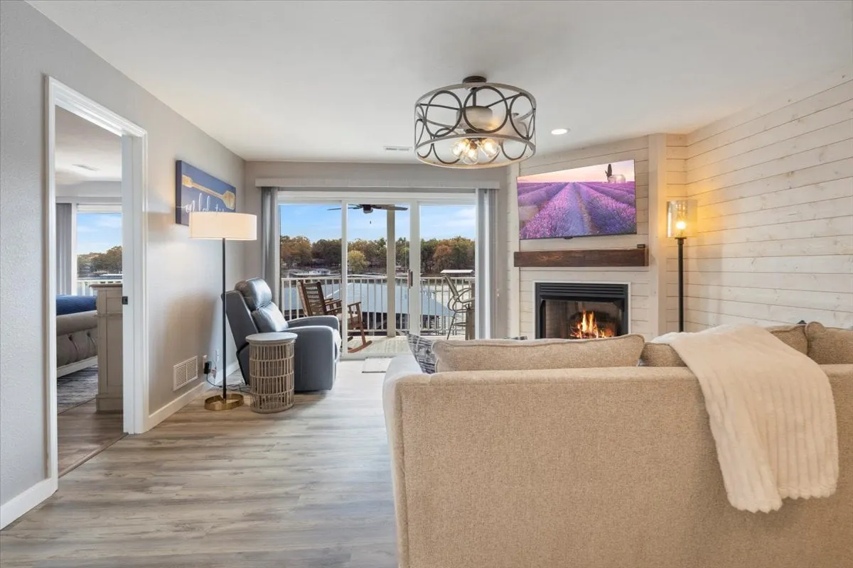 A bright, open living room with a contemporary gas fireplace as the centerpiece. Large, panoramic windows fill the space with natural light and offer stunning lake views. Plush, comfortable seating and cozy accents invite relaxation. A decorative light fixture and tasteful wall art add to the refined, welcoming atmosphere. The space is arranged for easy conversation and connection, with the view always in focus.