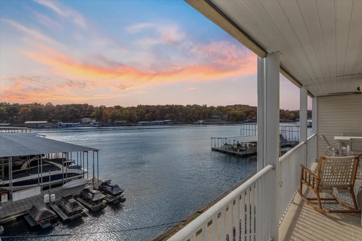 The image showcases an inviting lakeside porch with cozy rocking chairs overlooking calm waters, set against a stunning sunset sky. Boathouses dot the serene shoreline, and the golden hues reflect off the lake, creating a tranquil atmosphere ideal for relaxation or enjoying evening boat rides.