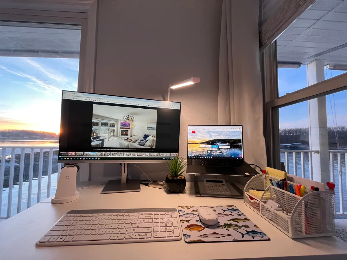 A dedicated workspace featuring computer monitors, a desk lamp, a plant, and colorful pens, all set up on a desk. The view from the desk shows the warm glow of sunset, creating an inspiring and tranquil atmosphere for remote work.