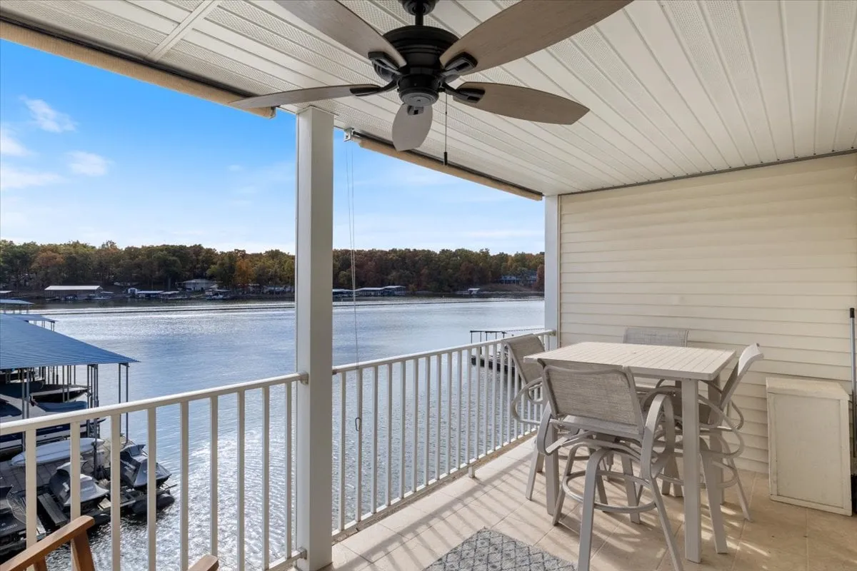 An exterior patio with an outdoor dining set overlooking the water. The tranquil scene includes a ceiling fan, lush greenery, and clear views of the lake, ideal for dining, relaxing, or enjoying sunset drinks.