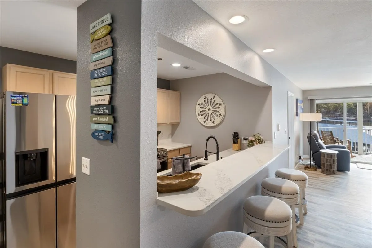 A modern kitchen with a spacious island, sleek stainless steel refrigerator, and bar stools arranged for casual gatherings. A decorative wall art piece adds personality, and the island is ready for meal prep or serving drinks. The angle also gives a glimpse of the patio outside, suggesting the lake's presence nearby and a transition between indoor and outdoor living.
